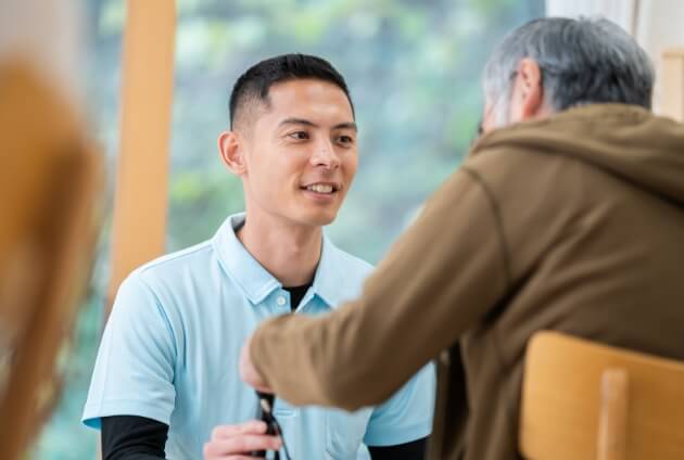 若い男性介護士が、高齢の利用者と向き合いながら会話をしている温かな場面